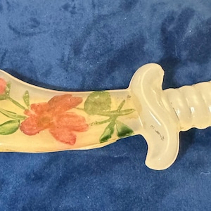 May include: A translucent, off-white dagger-shaped object made of a smooth material. The blade features a painted design of orange-red flowers with green leaves and stems. The handle has a ribbed, cylindrical texture with a curved crossguard.