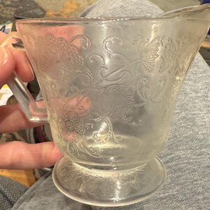 May include: A clear glass creamer with a handle and a spout. The creamer features an embossed floral pattern. The creamer is sitting on a gray surface.