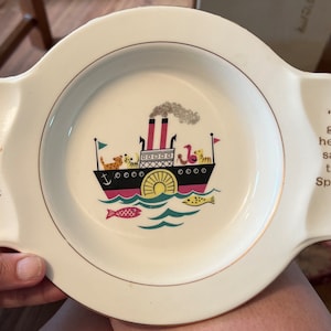 May include: A white ceramic children's bowl with a gold rim and a colorful illustration of a steamboat with animals on board. The text "I go here" says the Fork" and "I go here" says the Spoon" is printed on the sides of the bowl.