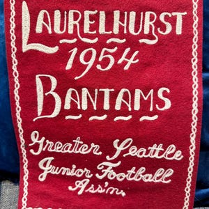 May include: A red felt pennant with white embroidered text. The text reads "LAURELHURST 1954 BANTAMS" and "Greater Seattle Junior Football Assn." A white stitched border frames the pennant.