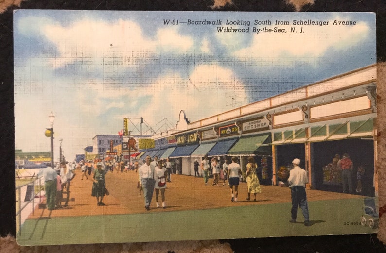 May include: A vintage postcard depicting a bustling boardwalk scene in Wildwood By-the-Sea, New Jersey. The postcard shows a view looking south from Schellenger Avenue, with shops and businesses lining the boardwalk. People are walking along the boardwalk, enjoying the summer day.