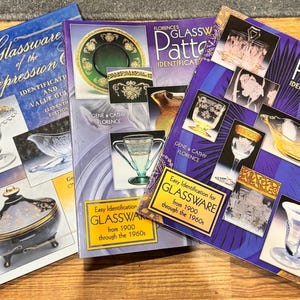 May include: Three books on glassware identification. The books are titled "Elegant Glassware of the Depression", and "Florence's Glassware Pattern Identification Guide" volumes I and IV. The books feature images of glassware and are on a wooden surface.