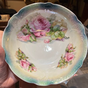 Puede incluir: Un tazón de porcelana blanca con un borde festoneado y un diseño floral con rosas rosadas y hojas verdes. El tazón tiene un esmalte iridiscente azul claro y verde.