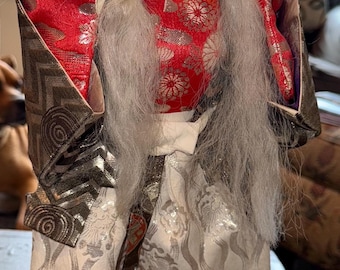 May include: A Japanese doll with a white face, red lips, and a stern expression. The doll has a large white wig and wears a red and gold patterned kimono with matching pants. The doll is standing on a black base.