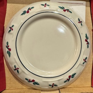 May include: Off-white ceramic plate with a dark blue border and a decorative holly pattern. The plate has a slightly raised rim with a woven texture and is nestled in a cardboard box. The holly design features green leaves and red berries.
