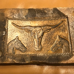 Puede incluir: Un molde rectangular de piedra gris con un diseño en relieve de una cabeza de toro y dos cabezas de caballo. El diseño está rodeado por un borde de cuerda.