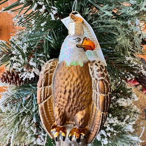 Puede incluir: Un adorno navideño de cristal con forma de águila calva, cuerpo marrón, cabeza blanca con purpurina iridiscente y pico naranja brillante. El águila está posada sobre una base gris texturizada con detalles de purpurina dorada en las alas.