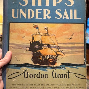 May include: A vintage book cover titled "SHIPS UNDER SAIL" by Gordon Grant. The cover features a detailed illustration of a sailing ship with orange sails against a sunset sky. The text below reads "THE SAILING VESSEL FROM THE EARLIEST TIMES TO THE PRESENT".
