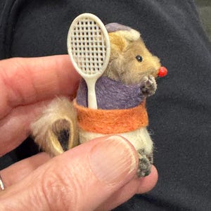 Puede incluir: Una figura de roedor en miniatura hecha a mano, con una raqueta de tenis. El roedor viste un atuendo morado y naranja, con una nariz roja. La figura se sostiene en una mano, mostrando los intrincados detalles del juguete.