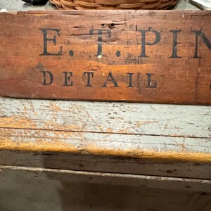 Op de afbeelding: Een verweerd houten bord met de tekst "E.T. PIN DETAIL" in zwarte letters. Het bord heeft een warme bruine kleur met zichtbare houtnerf en tekenen van slijtage. Een gevlochten mand en een afstandsbediening staan op de achtergrond.