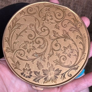 Op de afbeelding: Een rond, goudkleurig compact met een bloemenmotief. Het compact heeft een scharnierend deksel en een decoratief patroon van bladeren en bloemen.