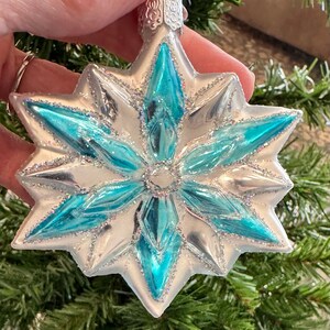 Op de afbeelding: Een zilveren en turquoise stervormige kerstversiering. Het ornament heeft een turquoise midden en zilveren randen met glitteraccenten. Het ornament wordt vastgehouden voor een groene kerstboom.