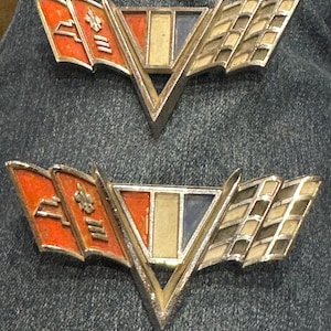 May include: Two vintage car emblems with a V-shaped design. Each emblem features a central section with red, white, and blue accents, flanked by orange flags and a checkered flag design. The emblems have a metallic finish.