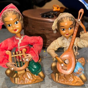 May include: Two vintage wooden dolls dressed in festive attire. One doll plays a harp, while the other plays a lute. Both dolls are sitting on wooden bases.