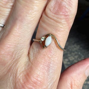 May include: A gold and silver ring featuring a white, oval-shaped stone and a small, clear gemstone. The ring has a distinctive, flowing design. The ring is on a finger.