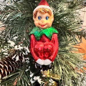 Könnte beinhalten: Ein Weihnachtsbaumschmuck aus Glas in Form eines Elfen mit roter Mütze, grünem Kragen und rotem Körper. Der Elf hat blaue Augen, braune Haare und ein lächelndes Gesicht. Der Schmuck ist mit Glitzer verziert und an einem goldenen Metallclip befestigt.