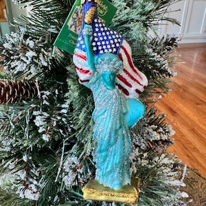 Könnte beinhalten: Ein Weihnachtsbaumschmuck aus Glas in Form der Freiheitsstatue, bestehend aus einer türkisfarbenen Figur mit silbernem Glitzer, die eine goldene Fackel und eine rot-weiß-blaue Flagge der Vereinigten Staaten hält, auf einem goldenen Sockel mit grünem Etikett.