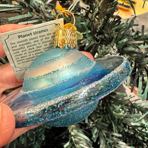 以下が含まれることがあります： 青と銀色のガラス製のクリスマスオーナメント。惑星の天王星の形をしており、キラキラと輝く氷のような外観で、金色の金属製のキャップが付いています。クリスマスツリーに飾られています。「Planet Uranus」と書かれたタグが付いています。