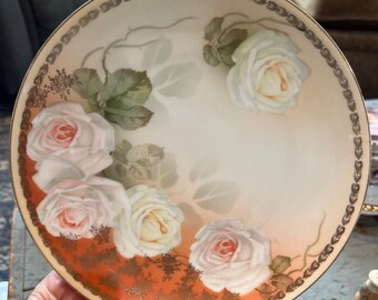 May include: A white porcelain plate with a gold rim and a floral design featuring white roses with orange accents. The plate has a vintage aesthetic.
