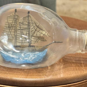 May include: A glass bottle with a miniature sailing ship inside. The ship is detailed with rigging and sails, set on a blue base. The bottle is sealed with a cork, and is resting on a wooden surface.