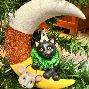 Puede incluir: Adorno navideño caprichoso con un gato negro con gorro de fiesta y cuello con volantes verdes, posado sobre una luna creciente amarilla. Un ratón gris está debajo. La luna tiene un acabado naranja y blanco brillante.