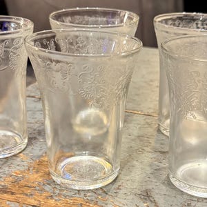 May include: A set of six clear, etched glass tumblers. Each glass features a flared shape and a decorative floral pattern near the rim. The glasses are arranged on a weathered wooden surface.