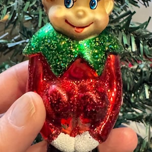 May include: A Christmas ornament shaped like an elf. The elf has a red glittery suit, a green glittery collar, and a red glittery hat with white trim. The elf's face is painted with blue eyes and a smiling expression. The ornament has a gold clip for hanging.