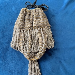 May include: A drawstring pouch covered in metallic silver and gold beads. The bag features a tiered design with long, dangling beaded fringe layers that extend from the main body, ending in a concentrated tassel at the bottom with black ties.