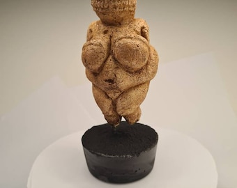Peut inclure: Une petite sculpture en céramique brune représentant une figure féminine aux traits exagérés. La sculpture est posée sur une base noire.