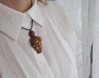Puede incluir: Un collar dorado con un colgante con forma de cuerpo de mujer. El colgante está detallado con una superficie texturizada y una pequeña cabeza.