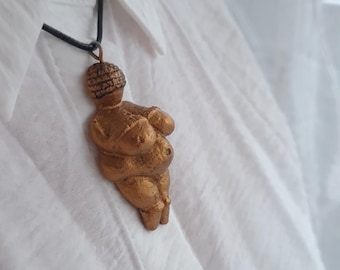 Puede incluir: Un collar con un colgante de color dorado en forma de figura femenina estilizada, con cabeza redonda y cuerpo completo. El colgante se lleva en un cordón negro.