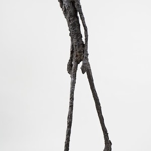 Può includere: Una scultura astratta nera di una persona che cammina. La scultura è realizzata in materiale ruvido e testurizzato e ha arti lunghi e sottili.