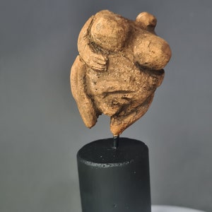 Può includere: Una piccola scultura in terracotta marrone di una persona che si tiene le braccia intorno a sé. La scultura è montata su un piedistallo nero.