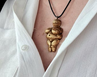 Peut inclure: Un collier avec un pendentif en forme de figurine féminine stylisée de couleur dorée, avec un corps rond et un visage simple. Le pendentif est porté sur un cordon noir.