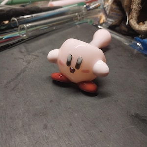 May include: A pink glass figurine of a cartoon character with a round body, small arms, and red feet. The character has black eyes and a smiling mouth. The figurine is on a dark gray surface.