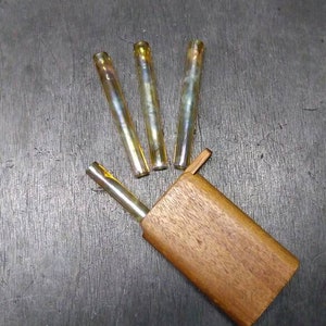 Könnte beinhalten: Ein Satz von vier gläsernen Rauchrohren mit einem Holzetui. Die Rohre sind irisierend und haben eine gelbe Tönung. Das Etui ist aus dunklem Holz.