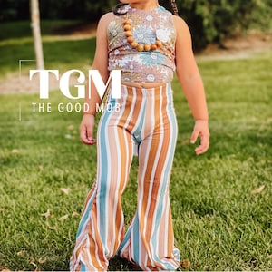 May include: A young child wearing a floral print halter top and striped bell-bottom pants. The outfit includes a matching headband with a large bow and a wooden bead necklace. The text "TGM THE GOOD MOB" is visible.