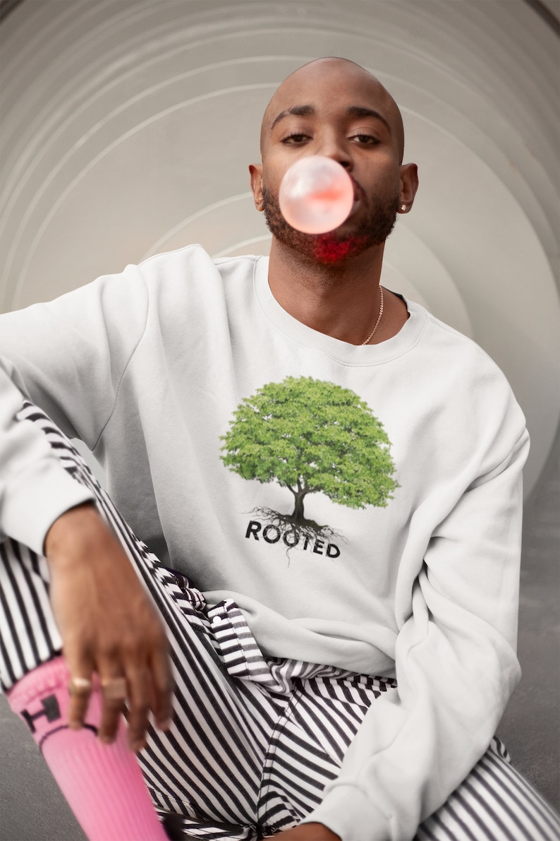 May include: A white sweatshirt with a green tree graphic and the word "ROOTED" printed below it. The person wearing the sweatshirt is blowing a bubble with pink bubble gum.