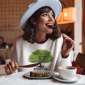 May include: A woman wearing a white sweatshirt with a green tree graphic and the word "ROOTED" printed below it. She is sitting at a table in a cafe and eating a piece of cake.