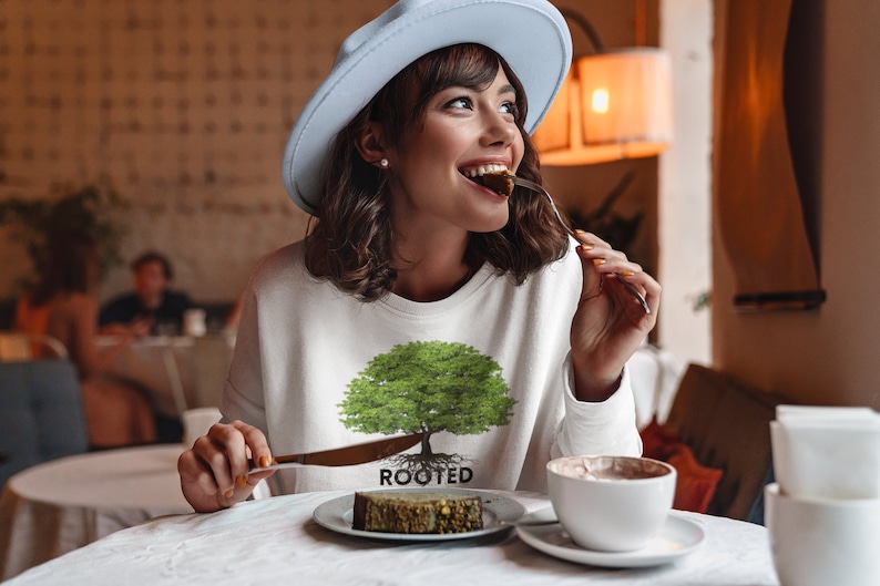May include: A woman wearing a white sweatshirt with a green tree graphic and the word "ROOTED" printed below it. She is sitting at a table in a cafe and eating a piece of cake.