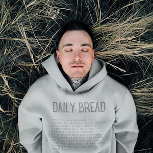 Może przedstawiać: Osoba w szarej bluzie z kapturem z napisem "DAILY BREAD" i nadrukowaną modlitwą z przodu. Osoba leży na plecach na polu wysokiej trawy.