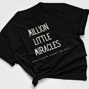 Può includere: T-shirt nera con testo bianco che recita "MILLION LITTLE MIRACLES" impilato verticalmente. Sotto, la frase "I CAN'T EVEN COUNT'EM ALL!" è stampata. La maglietta è annodata in basso.