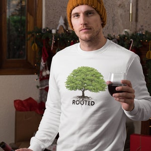 May include: A white sweatshirt with a green tree and the word "ROOTED" printed on the front. The person wearing the sweatshirt is holding a glass of red wine and a bottle of wine.