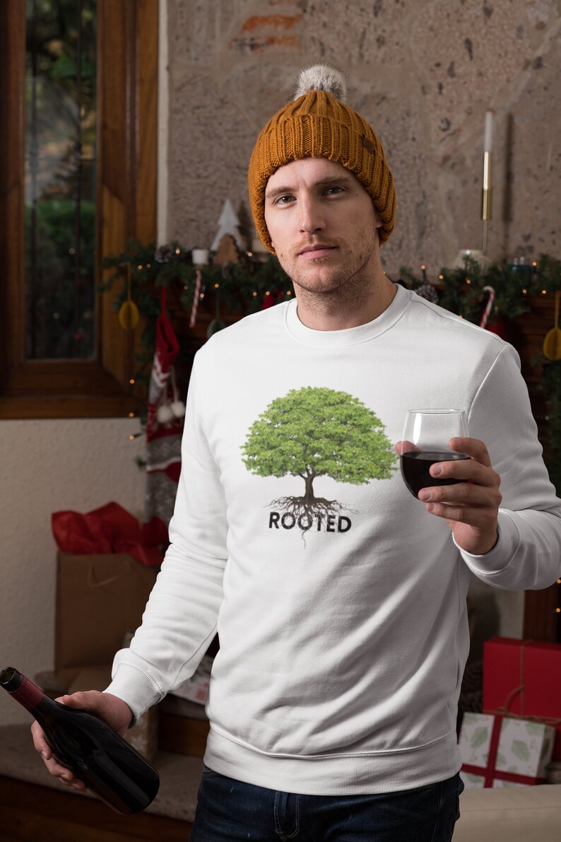 May include: A white sweatshirt with a green tree and the word "ROOTED" printed on the front. The person wearing the sweatshirt is holding a glass of red wine and a bottle of wine.