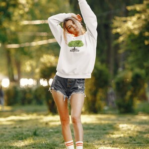 May include: A white sweatshirt with a green tree graphic and the word "ROOTED" printed below it. The model is wearing denim shorts and white socks with orange stripes.