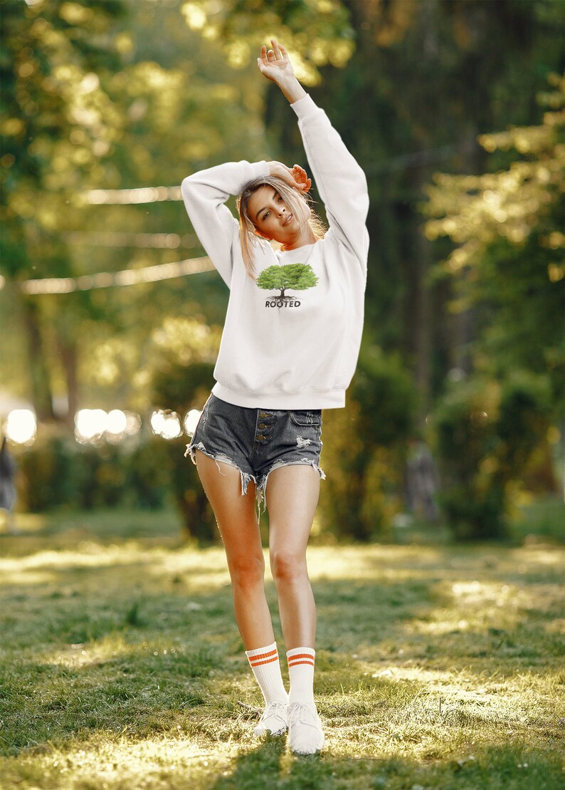 May include: A white sweatshirt with a green tree graphic and the word "ROOTED" printed below it. The model is wearing denim shorts and white socks with orange stripes.