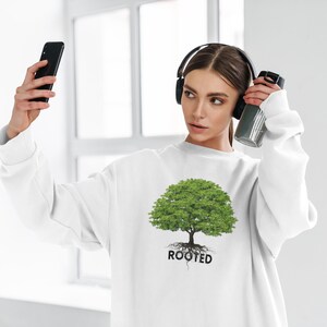 May include: White sweatshirt with a green tree graphic and the word "ROOTED" printed below. The person is wearing headphones and holding a water bottle.