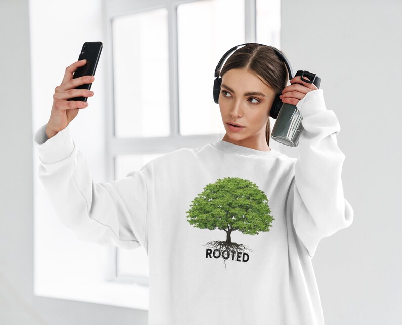 May include: White sweatshirt with a green tree graphic and the word "ROOTED" printed below. The person is wearing headphones and holding a water bottle.