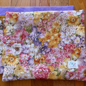 May include: A quilted floral laptop sleeve with a vibrant pattern of pink, yellow, and purple flowers. The sleeve is layered over a solid lavender fabric. A gold zipper and charm are visible on the side. The label reads "Stars & Stitches by Anna B."