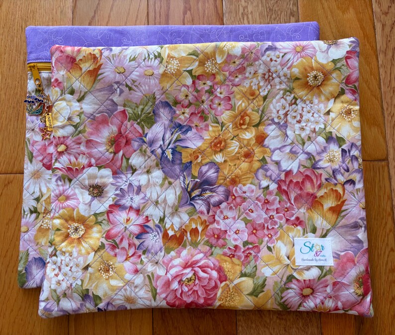 May include: A quilted floral laptop sleeve with a vibrant pattern of pink, yellow, and purple flowers. The sleeve is layered over a solid lavender fabric. A gold zipper and charm are visible on the side. The label reads "Stars & Stitches by Anna B."